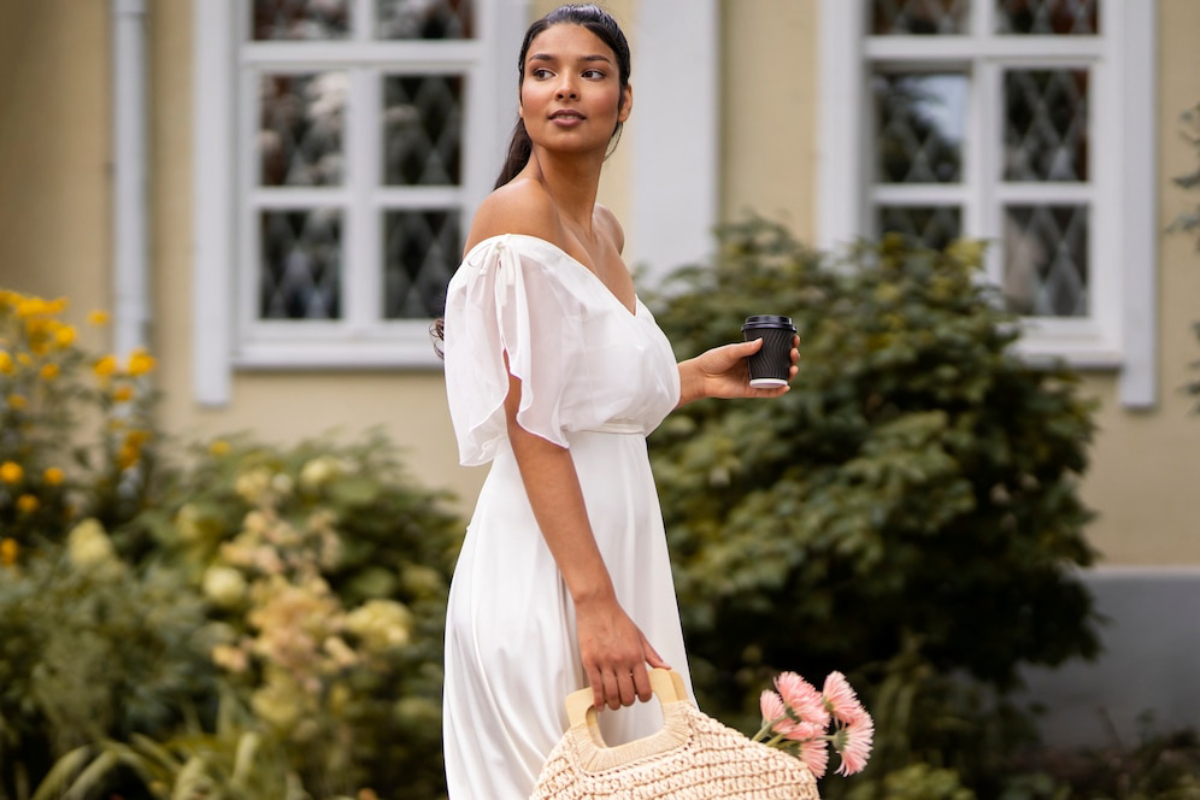 A woman in a white dress walks outside, holding a coffee cup and a woven bag with flowers, surrounded by green foliage and a house.