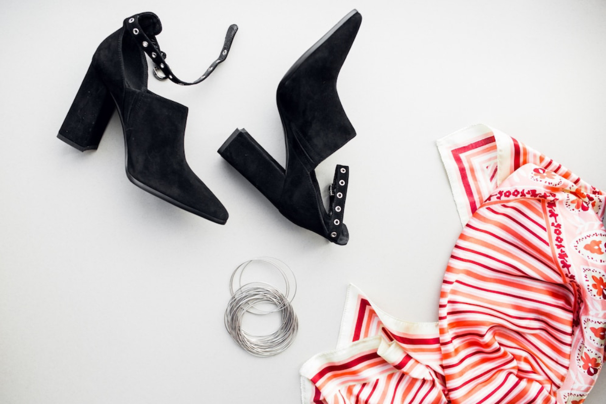 Pair of black suede high-heeled shoes with ankle straps, silver bangles, and a patterned red and white scarf on a gray background.
