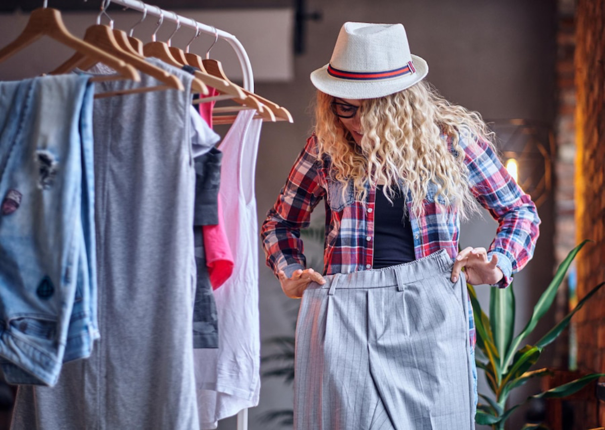 A woman with curly hair and glasses tries on a pair of gray striped pants while standing in a stylish clothing store.