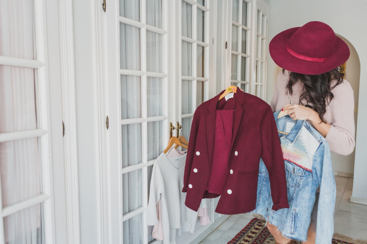 A woman in a cozy room holds a burgundy blazer and a denim jacket.