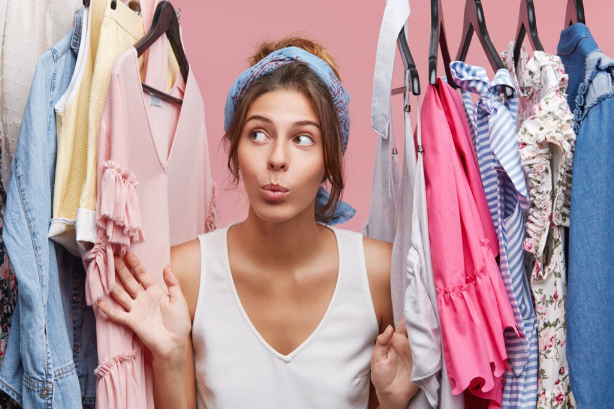 A woman wearing a white tank top stands between colorful dresses on hangers against a pink background.