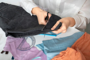 A person cuts the hem of dark jeans with scissors, surrounded by colorful fabric scraps and a protractor on a table.