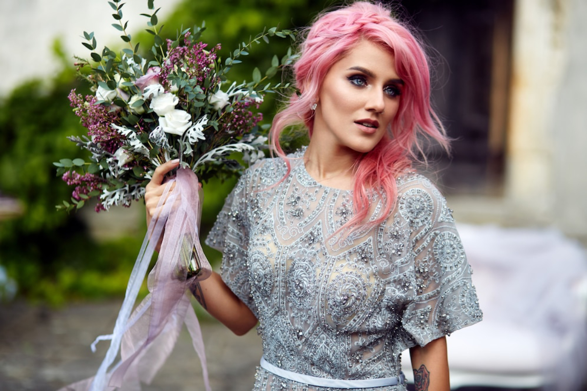 A person with pink hair holds a lush bouquet of white and purple flowers, wearing a sparkling silver dress.