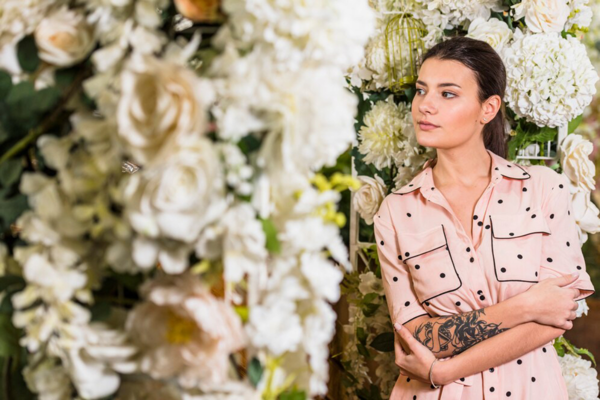 A woman in a pink polka-dot shirt stands among a lush backdrop of white and pastel flowers.