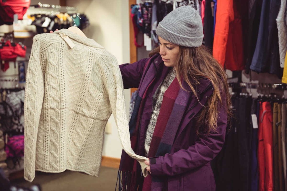 A person in a purple coat holds a cream-colored cable-knit sweater in a clothing store surrounded by various colorful garments.