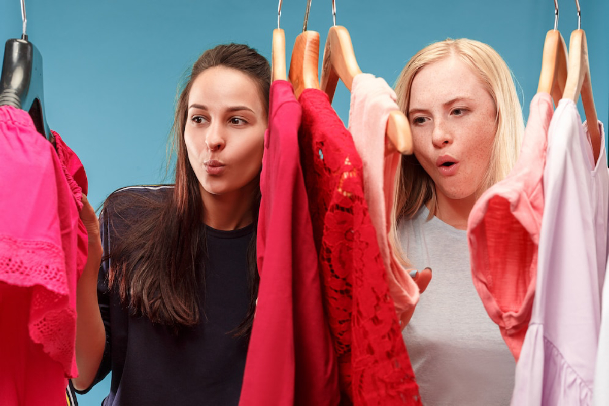 Two women select clothing from a vibrant rack filled with pink, red, and white garments against a light blue background.