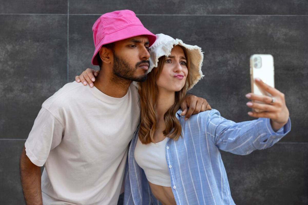 A couple wearing colorful hats takes a selfie together, leaning in close, against a modern gray background.