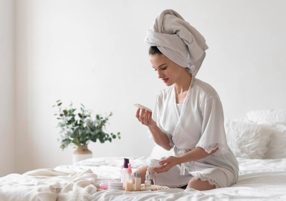 A woman in a white robe with a towel turban sits on a bed, surrounded by skincare products and a small plant.