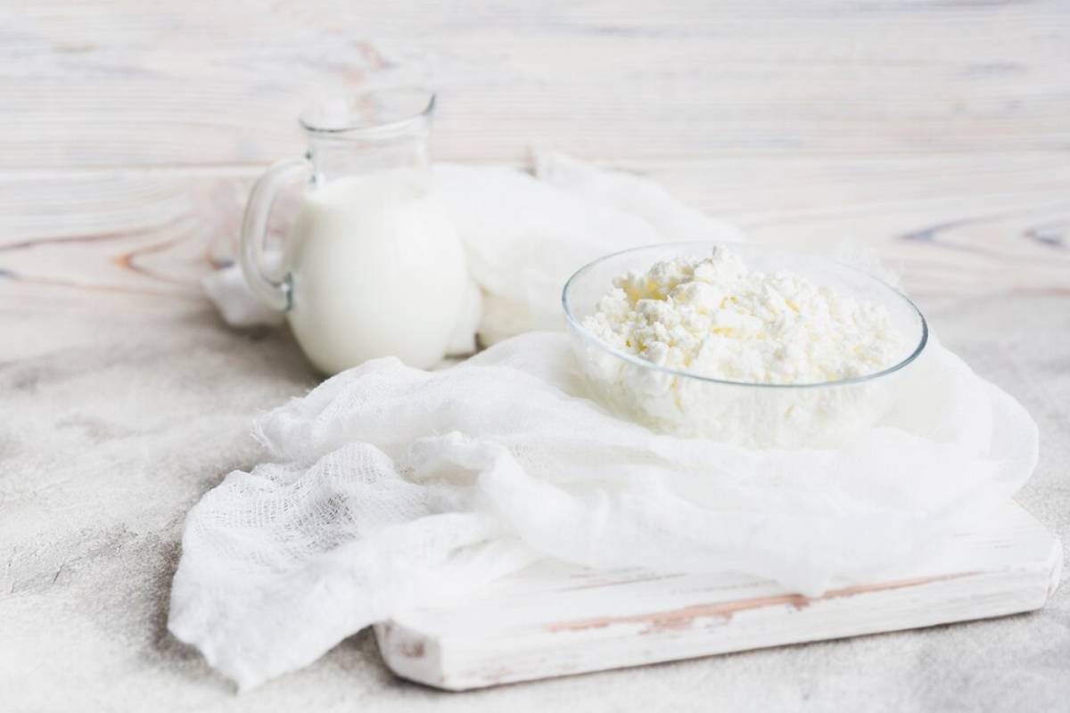 A clear bowl of cottage cheese next to a jar of milk, both placed on a white cloth over a rustic wooden surface.