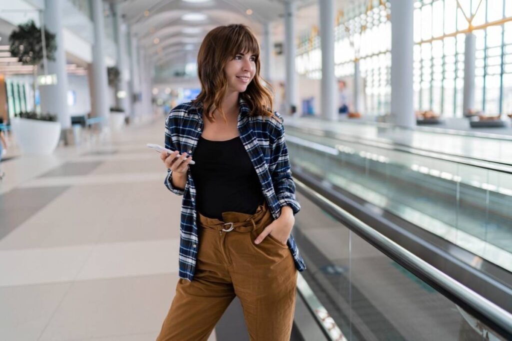 Young woman holding a smartphone in an airport terminal, smiling and looking to the side.